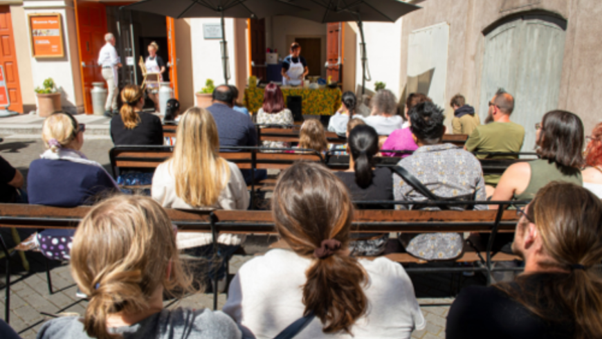 People sitting in the sun watching a person in an apron give a demonstration