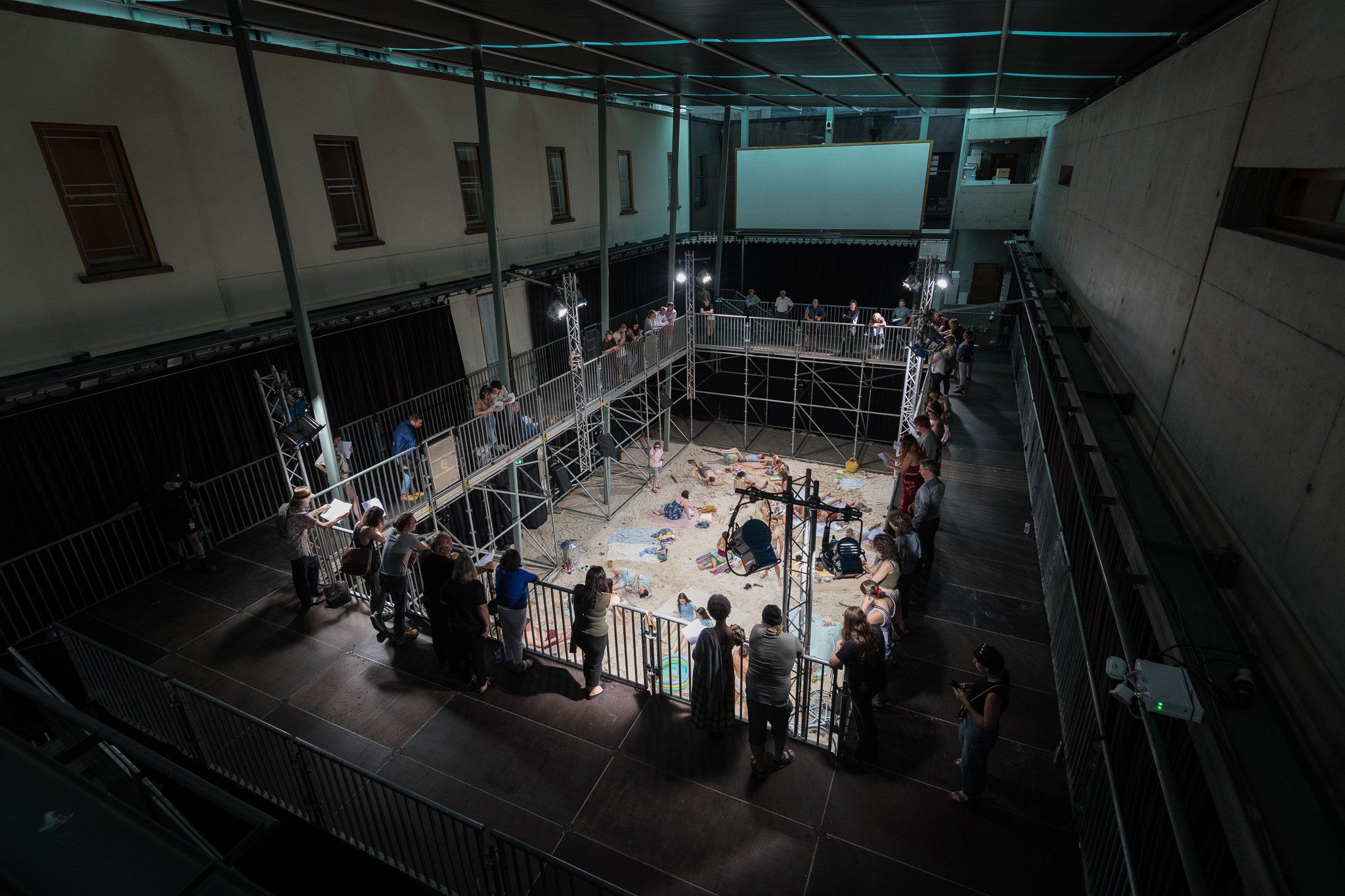 A beach is created inside Millennium Hall, using sand. The sand is surrounded by scaffolding. On the scaffolding an audience watches as performers put on an opera